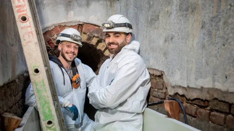 Renovatiewerf in een smalle ondergrondse rioolruimte, waar twee personen in witte beschermende pakken en helmen staan voor een opengelegde bakstenen wandboog. De omgeving toont ruwe muren, bloot metselwerk en werfmateriaal.