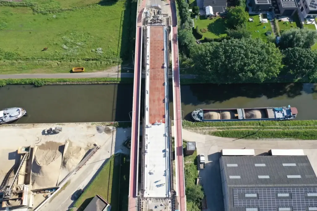Vue aérienne d’un pont en rénovation au-dessus d’un canal, avec des matériaux de chantier et des zones de travail sur le tablier du pont. Des bateaux sont amarrés de chaque côté de l’eau, entourés de verdure et de bâtiments industriels à proximité.
