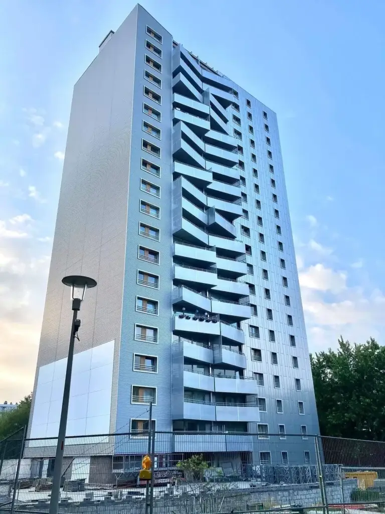 Immeuble résidentiel de grande hauteur avec une façade rénovée et des balcons originaux en forme de zigzag, photographié depuis le niveau de la rue. Le bâtiment présente une finition moderne de couleur argentée et se trouve à côté d’une zone de chantier clôturée avec du matériel et des barrières.