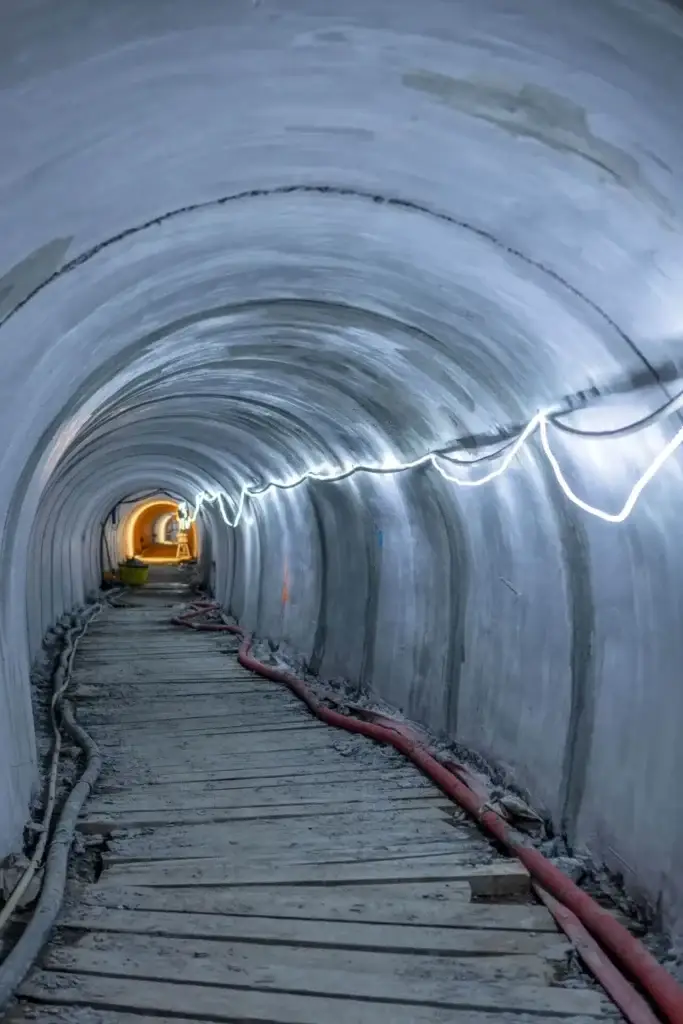 Tunnel d’égout souterrain avec un plancher de circulation en bois, des conduites rouges et grises le long des parois et une rangée d’éclairages de chantier lumineux éclairant la structure en béton courbée. Un éclairage supplémentaire est visible au bout du tunnel.