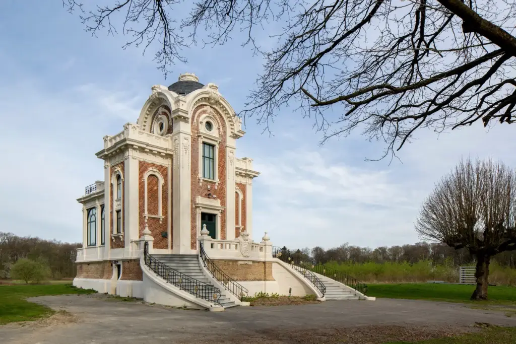 Bâtiment historique avec un escalier monumental et une façade richement travaillée, entouré d’un paysage verdoyant ouvert et d’arbres. Architecture symétrique avec des éléments décoratifs et de grandes fenêtres.