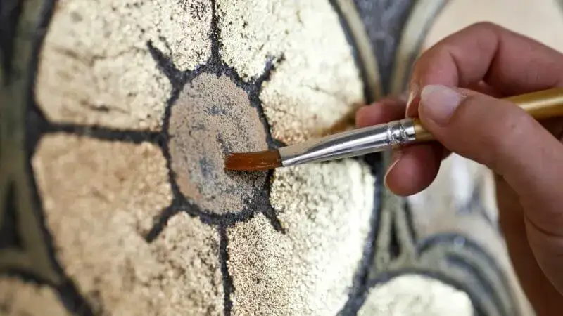 Close‑up van een restaurator die met een fijn penseel goudkleurige details op een historisch muurschilderingsornament retoucheert, met zicht op textuur, pigmenten en decoratieve patronen.