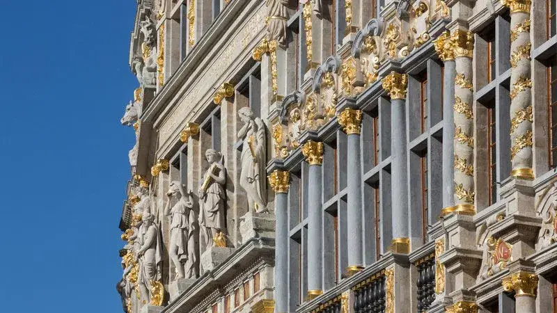 Detailbeeld van een rijk gedecoreerde historische gevel met sculpturen, zuilen en vergulde ornamenten. De natuurstenen elementen en gouden accenten worden duidelijk uitgelicht tegen een heldere blauwe lucht.