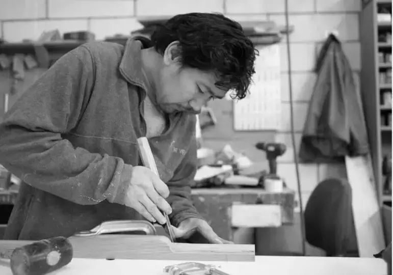 Ambachtelijke houtbewerker die in een werkatelier met handgereedschap fijn schrijnwerk uitvoert, omringd door timmermateriaal en een traditionele werkbank.