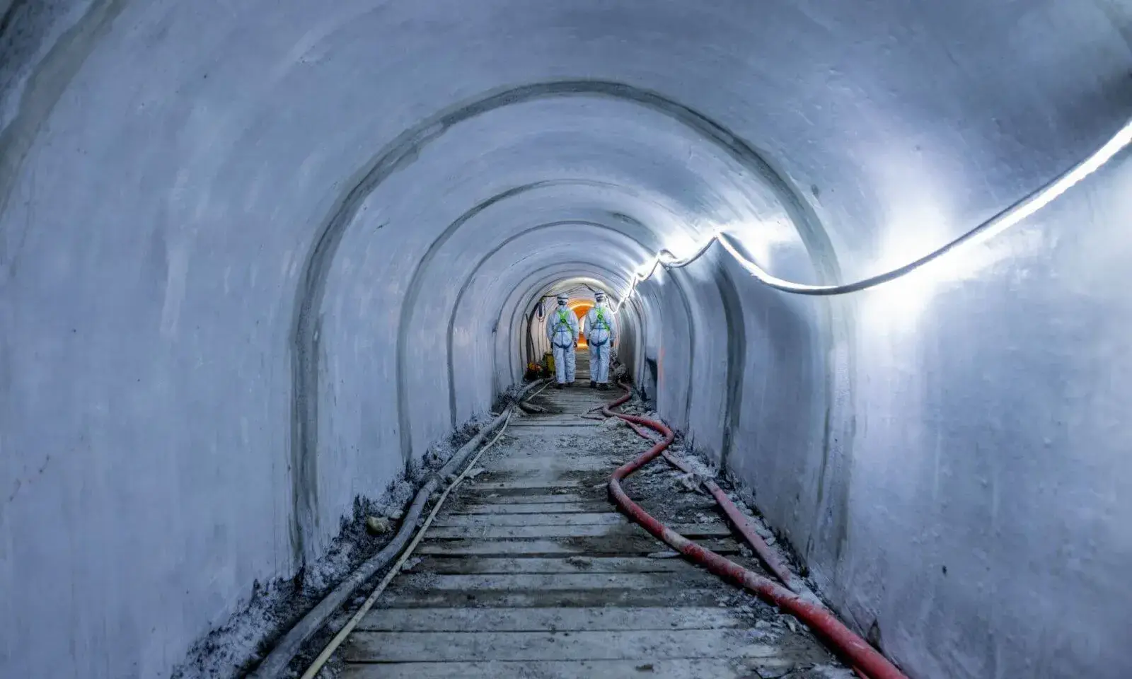 Ondergrondse riooltunnel waar twee medewerkers in beschermende kledij aan het einde van de doorgang staan tussen werkleidingen en een houten loopvloer tijdens rioolrenovatiewerken.