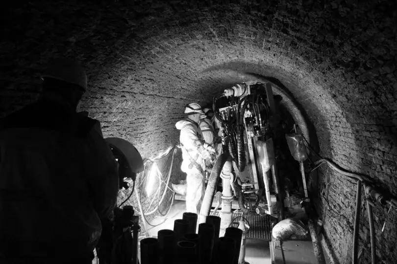 Technisch team voert groutpalen‑ of injectiewerken uit in een ondergrondse gemetselde tunnel, met gespecialiseerde boor‑ en injectiemachines, verlichting langs de wand en verschillende werkleidingen en slangen.