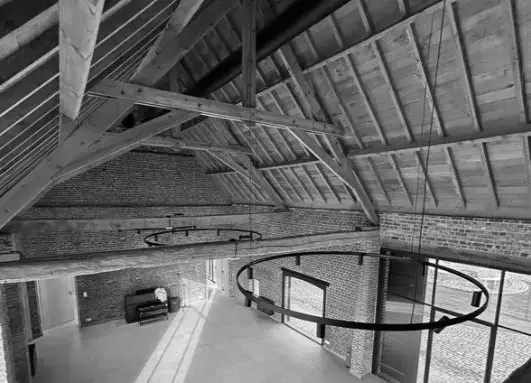 Gerestaureerde erfgoedruimte met open zadeldak, zichtbare houten spanten, bakstenen muren en grote moderne raamopeningen, als onderdeel van een herbestemmings‑ en renovatieproject.