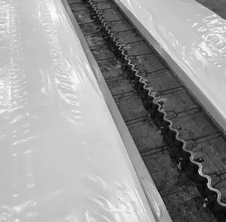 Detail van een betonnen vloer met openliggende voegconstructie waarin een metalen profiel zichtbaar is, geflankeerd door twee breed aangebrachte waterdichtings‑ of kunststofmembranen, als onderdeel van voegafdichtings‑ en vloerrenovatiewerken.