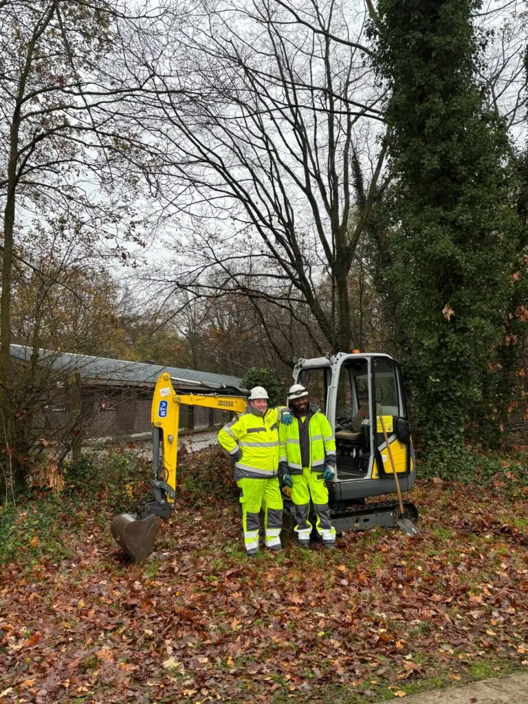 Twee personen in fluogele werkkledij en veiligheidshelmen staan buiten naast een kleine graafmachine op een terrein met herfstbladeren, met bomen en een gebouw op de achtergrond.