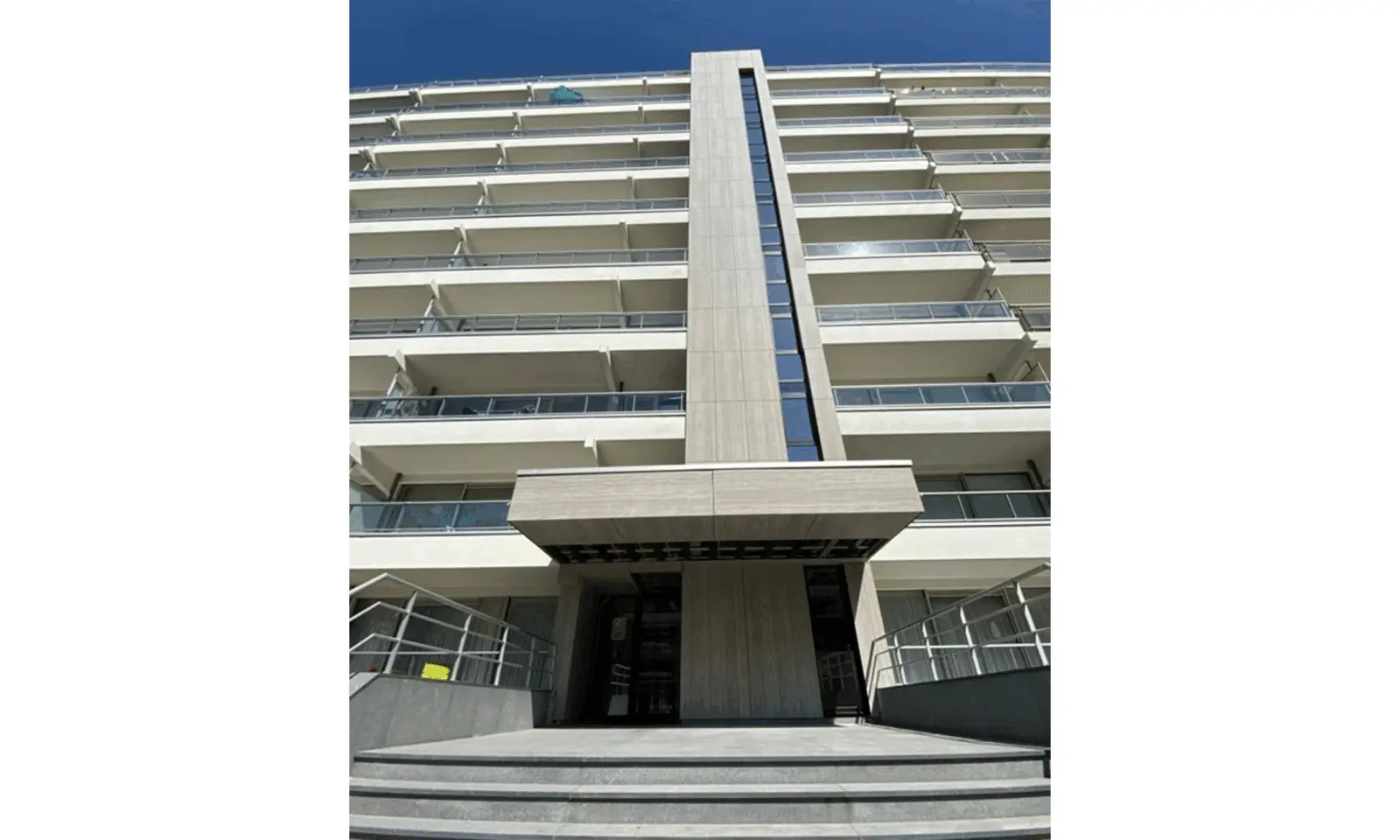 Een modern gebouw met meerdere verdiepingen en glazen balkons, gezien vanaf de onderkant. De ingang heeft brede treden die leiden naar een hoog verticaal architectonisch element boven de deuropening.