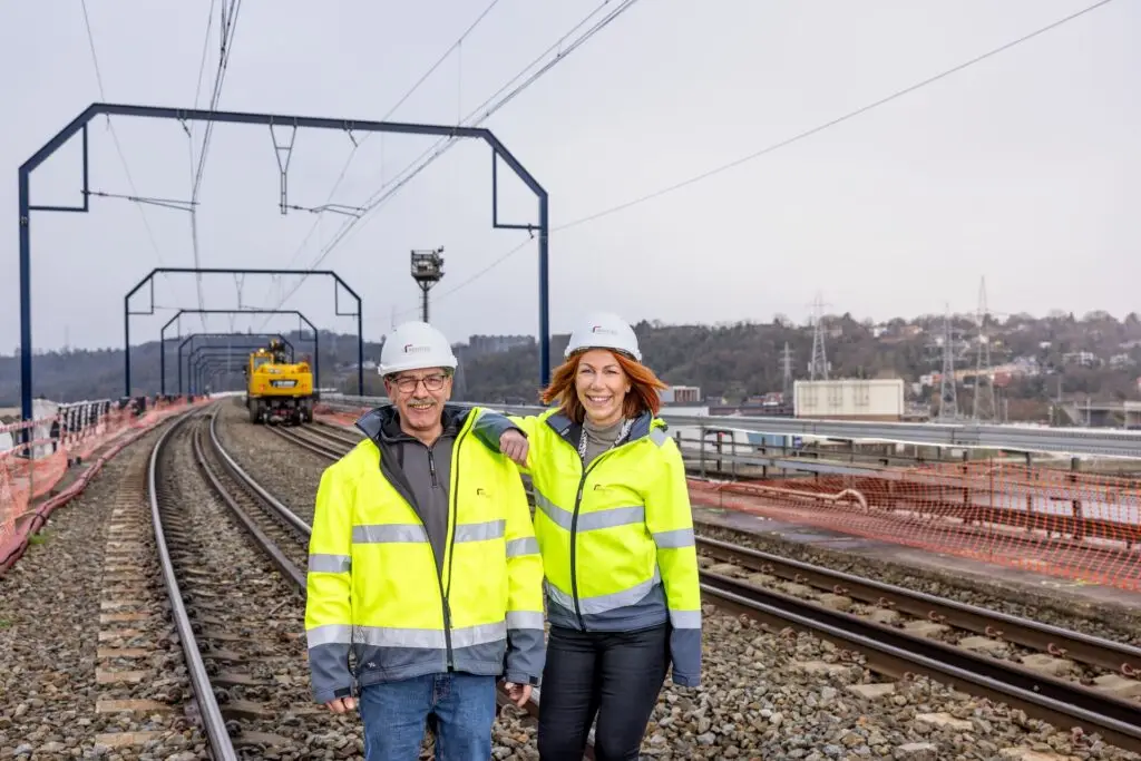 Twee Renotec-medewerkers in signalisatiekledij op spoorwerf met bovenleiding en spoorinfrastructuur