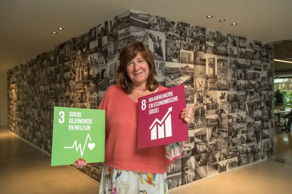 CFO van Renotec houdt SDG-borden ‘Goede gezondheid en welzijn’ en ‘Waardig werk en economische groei’ vast in kantoorruimte met referenties naar renovatieprojecten