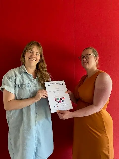 Twee personen houden officieel certificaat of attest vast tegen rode achtergrond, overhandiging door externe organisatie als erkenning voor inclusieve werkcultuur en gelijke kansen binnen Renotec