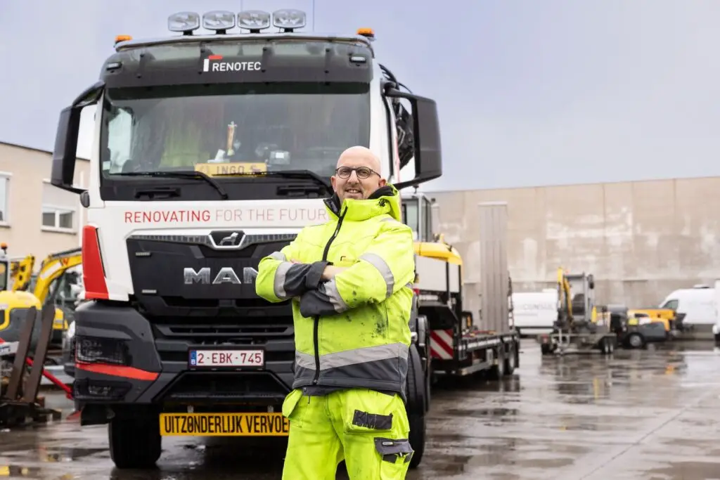 Renotec chauffeur in fluorescerende werkkledij staat op een werf voor een Renotec-vrachtwagen met kraan, klaar voor transport en logistieke ondersteuning van bouwmaterialen op de werf.